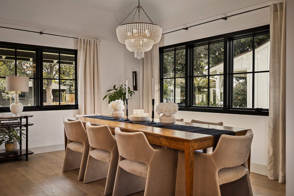 Dining room with an oak table, velvet chairs, and a beaded chandelier between black framed windows