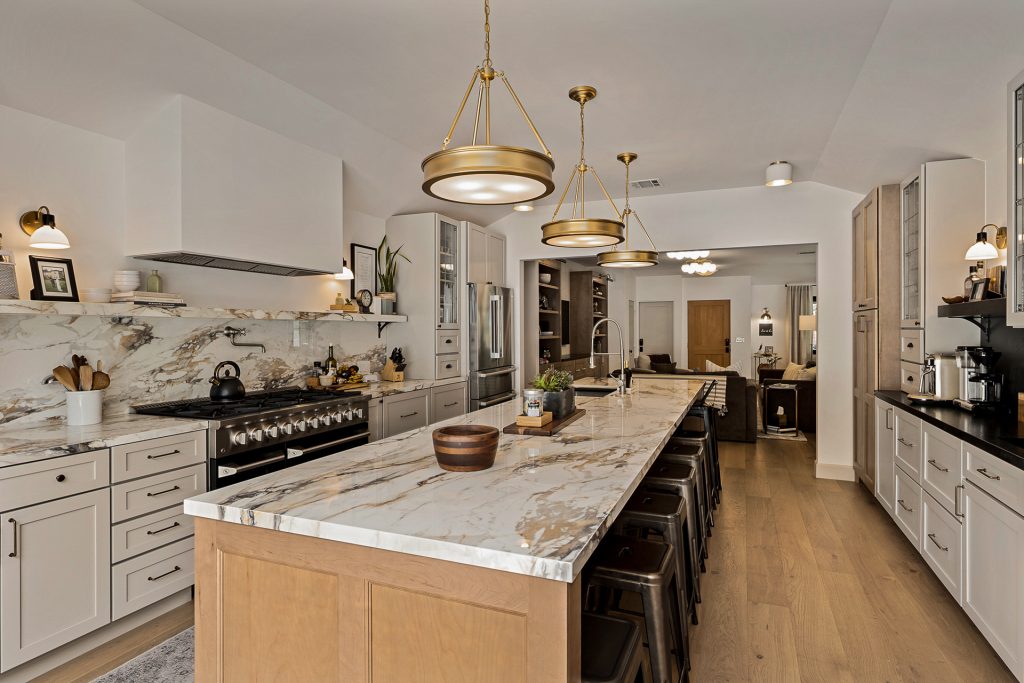 Kitchen island topped in veined marble under three brass drum pendants, with the range and marble slab backsplash beyond