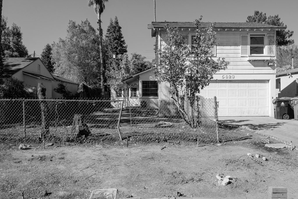 Black and white photo of the original two-story house before renovation, with bare dirt yard and chain-link fence