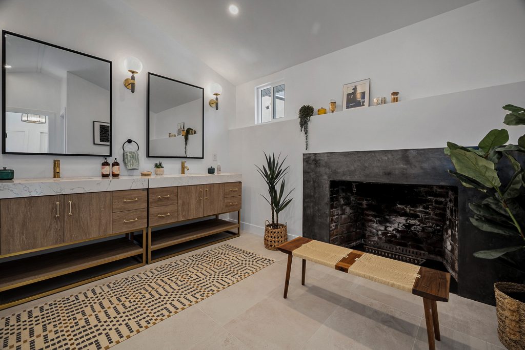 Primary bathroom with a blackened plaster fireplace, oak double vanity on a brass base, and a woven bench
