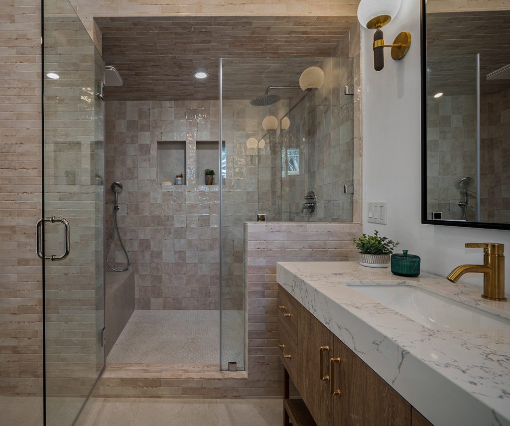 Primary bathroom walk-in shower in stone tile behind a glass enclosure, next to a marble topped oak vanity