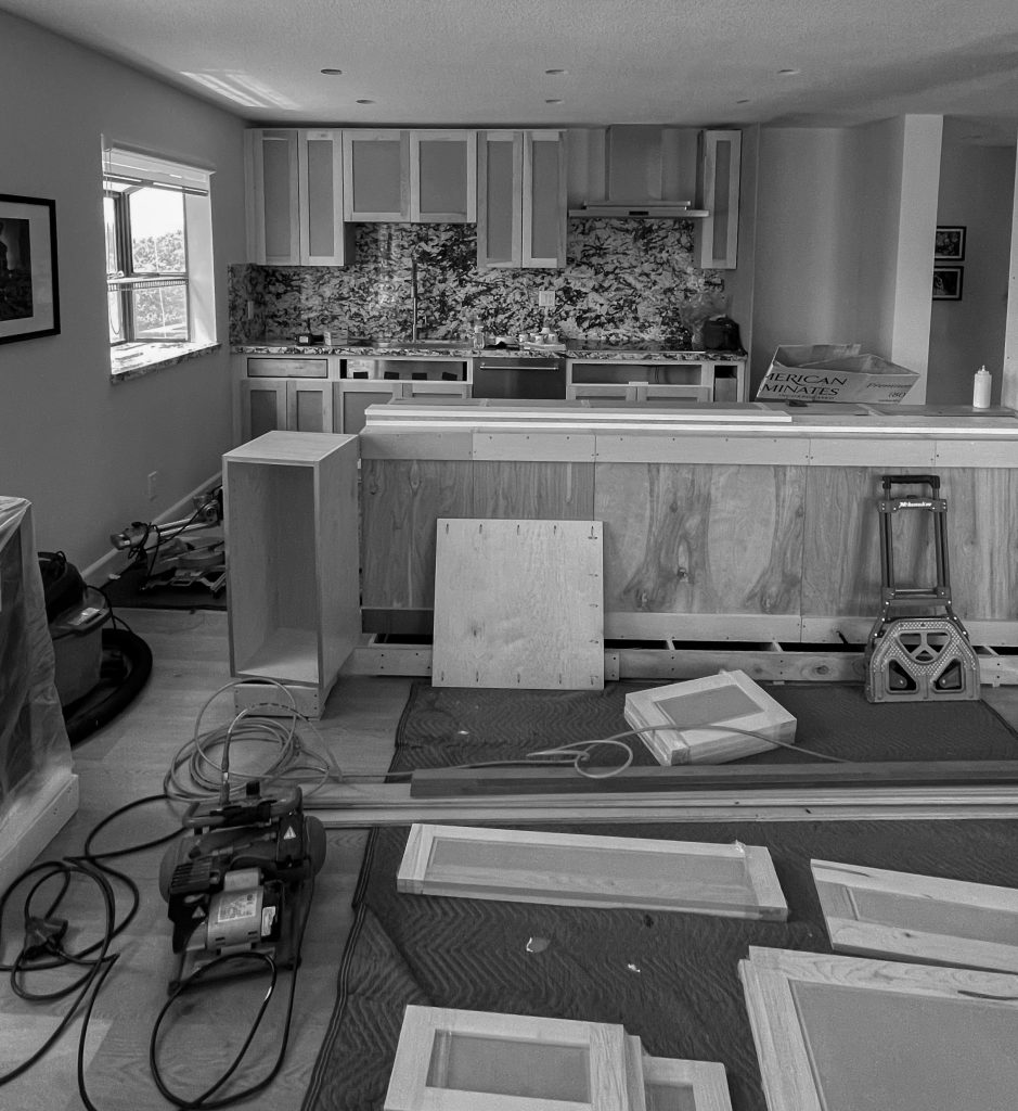 Kitchen during construction, with the island framed in plywood, upper cabinets hung, and cabinet doors laid out across the floor