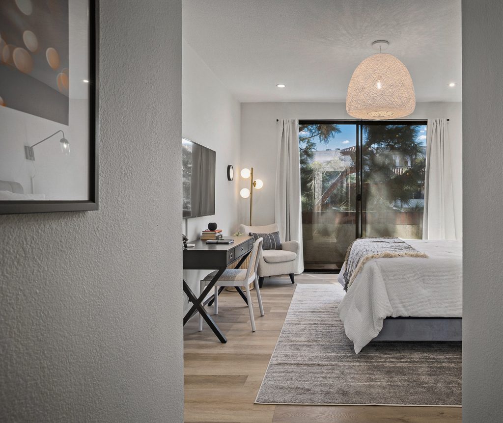 Guest bedroom seen through the doorway, with a woven pendant and a desk beside the balcony door