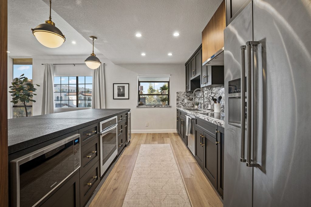 Galley run of the kitchen with dark cabinetry, wall ovens, and granite carried up the backsplash