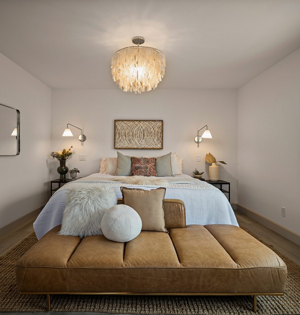 Primary bedroom with a capiz shell chandelier, brass swing arm sconces, and a tan leather bench at the foot of the bed