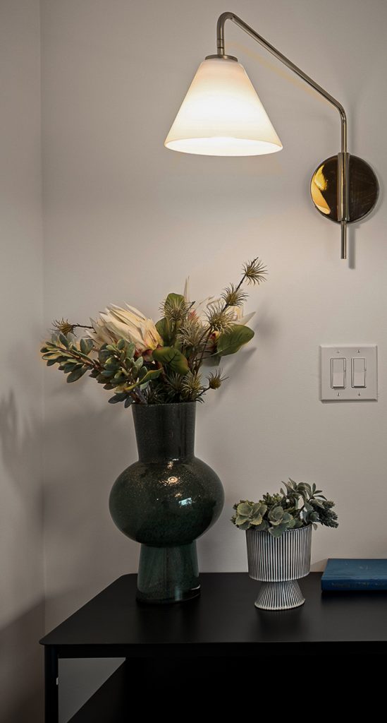 Brass swing arm sconce lighting a black console styled with a dark green glass vase