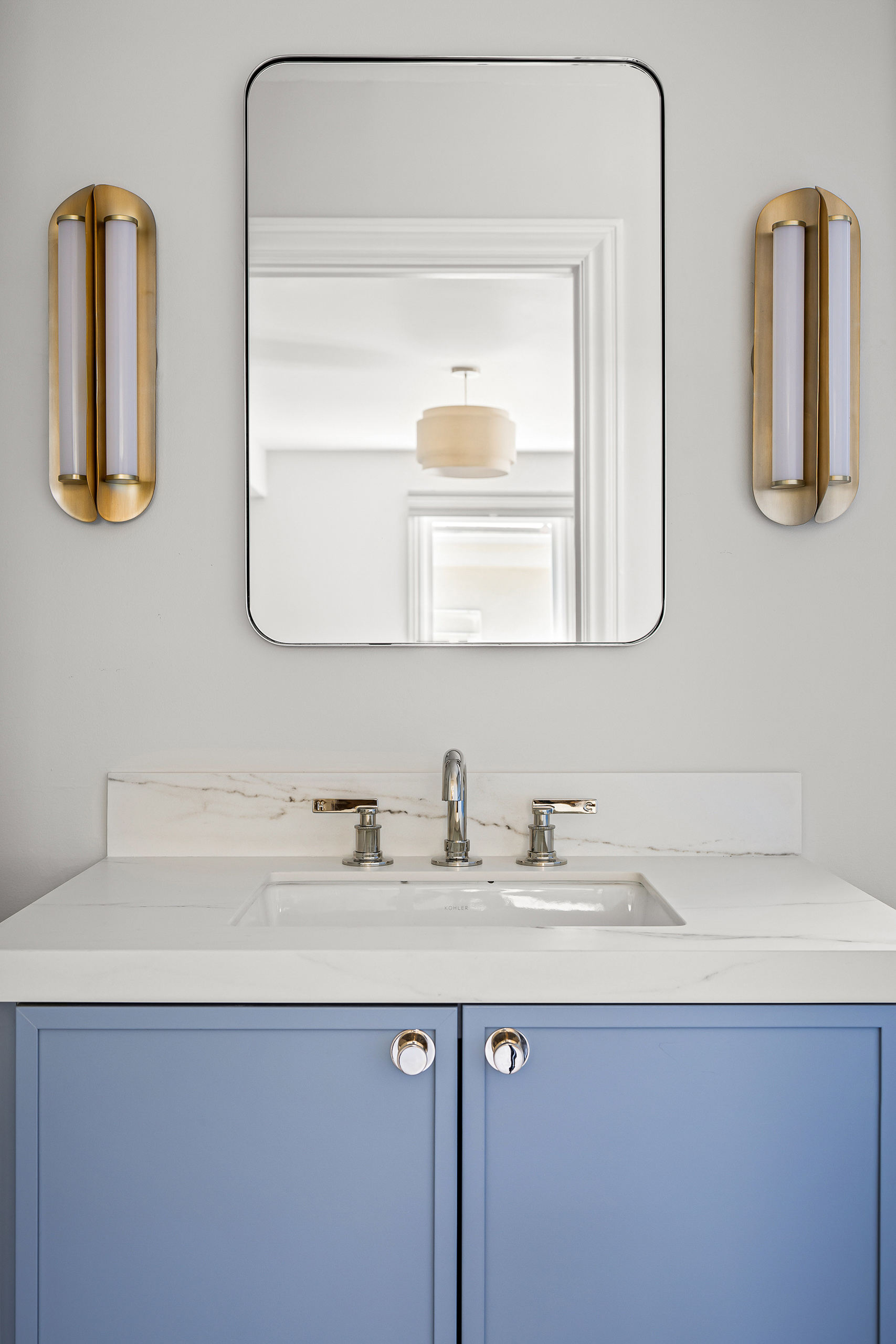 The finished vanity in soft blue with a marble top, a rounded rectangular mirror and a brass sconce on either side.