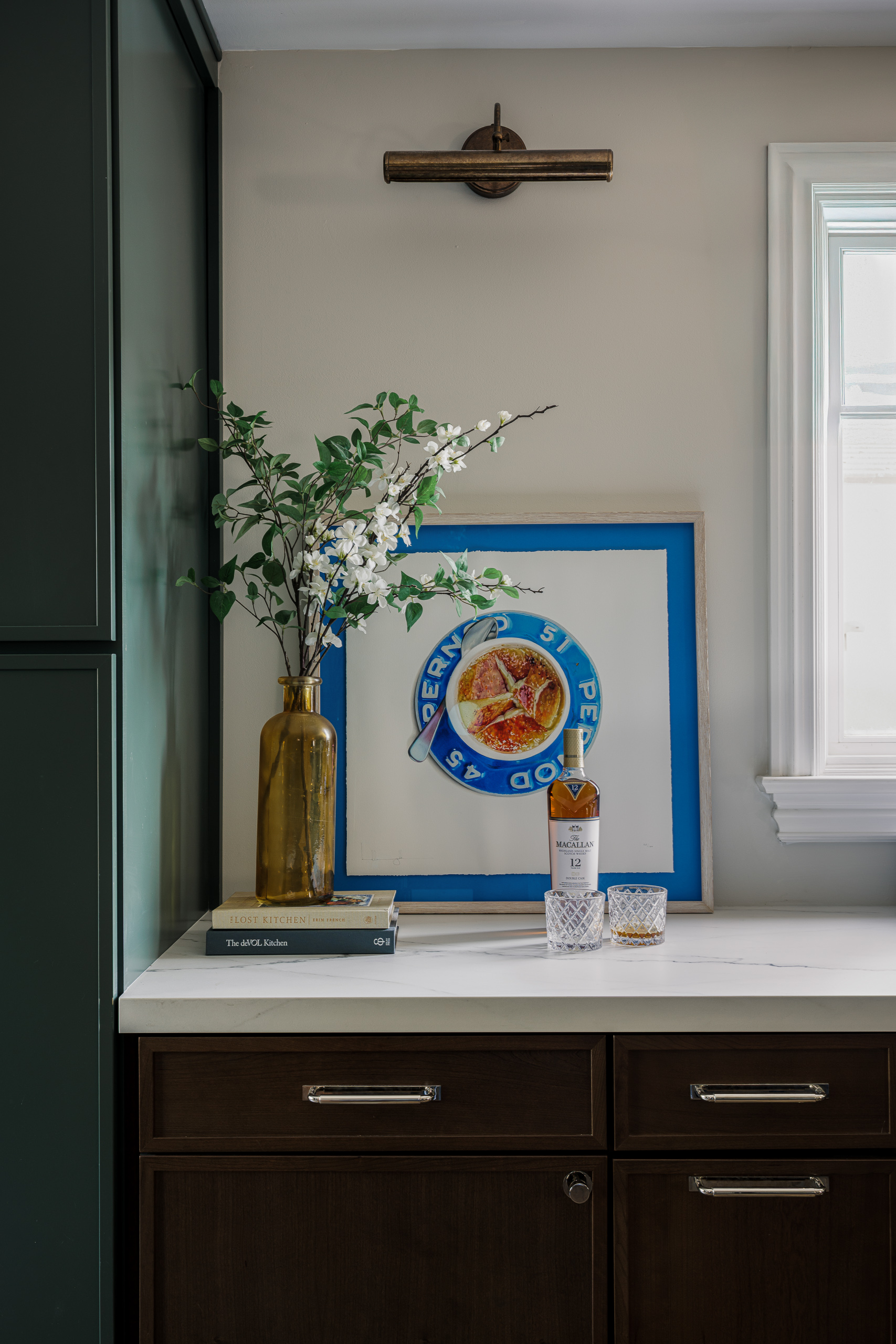 The coffee bar counter with a framed print leaning against the wall beneath a brass picture light, next to a tall green cabinet.
