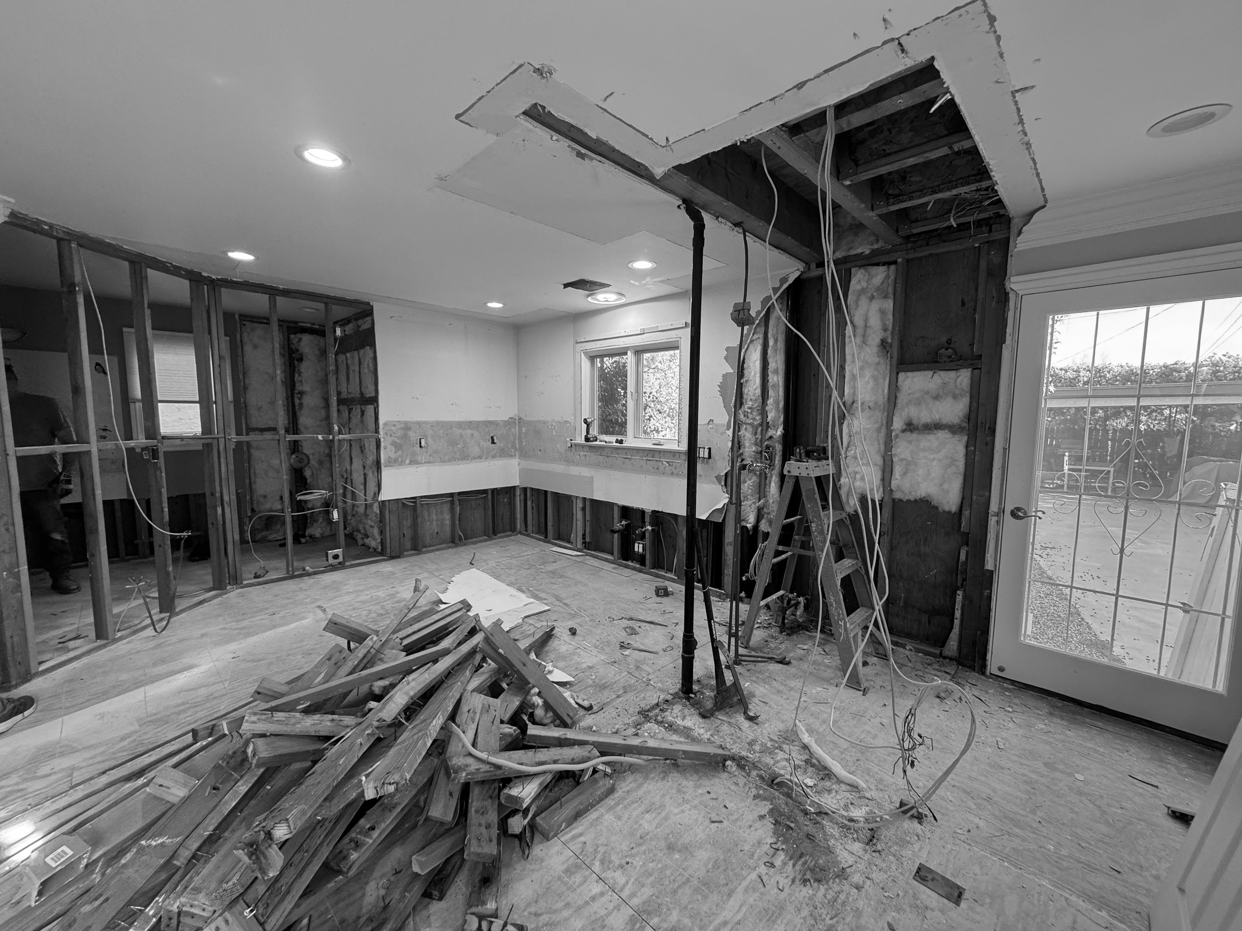 The kitchen during construction with the ceiling opened, a temporary support post in place, framing lumber stacked on the floor and the French door opening at right.