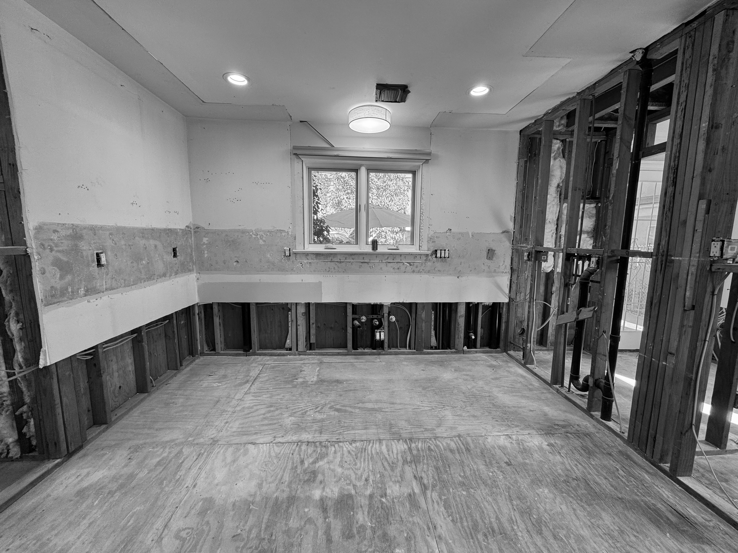 The kitchen during construction, drywall cut away in a straight line around the room, exposed studs and plumbing at the sink wall below the window.