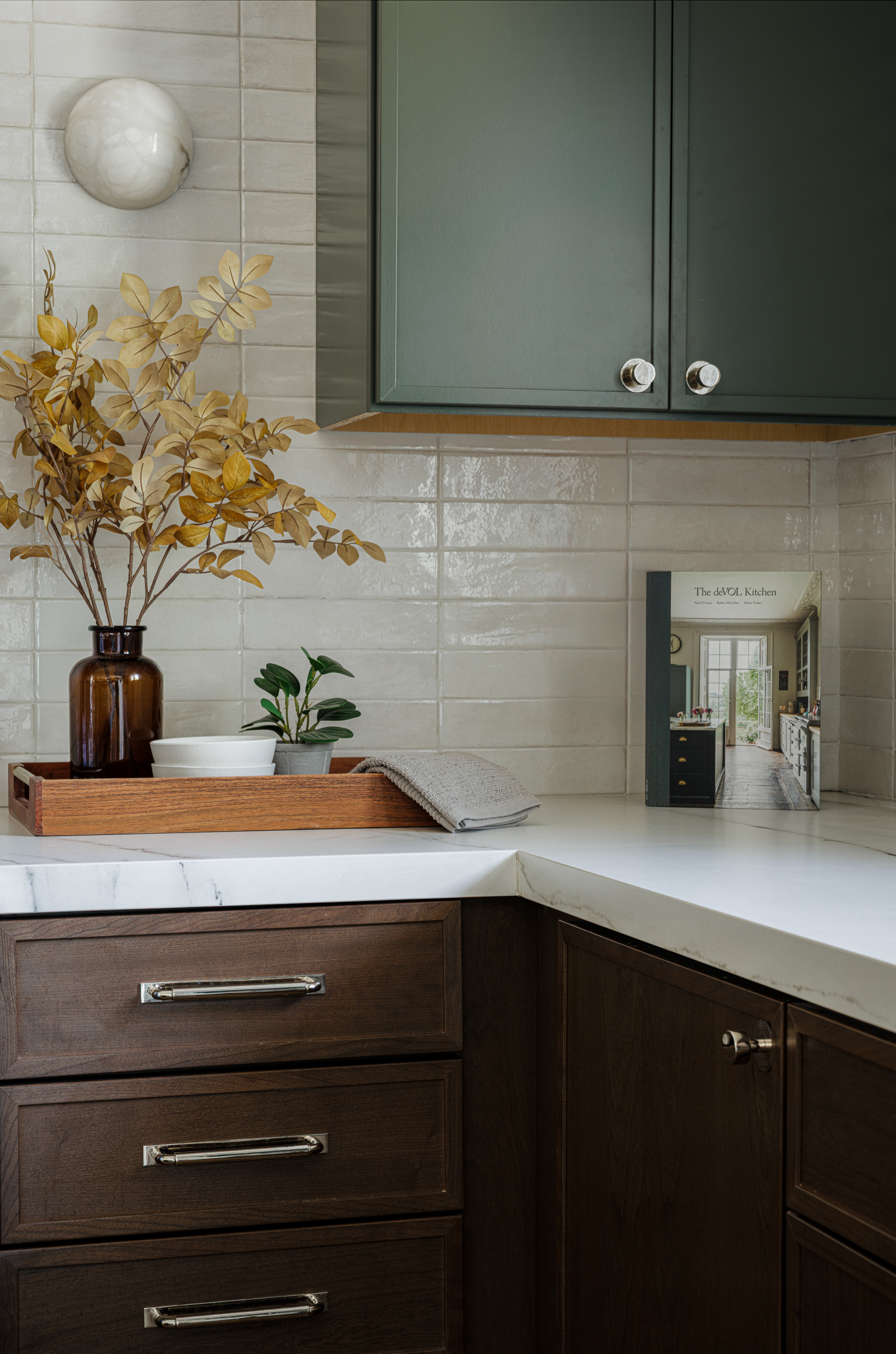 A corner of the kitchen counters, dark wood drawers with polished nickel cup pulls beneath a marble top holding a wooden tray and a cookbook.