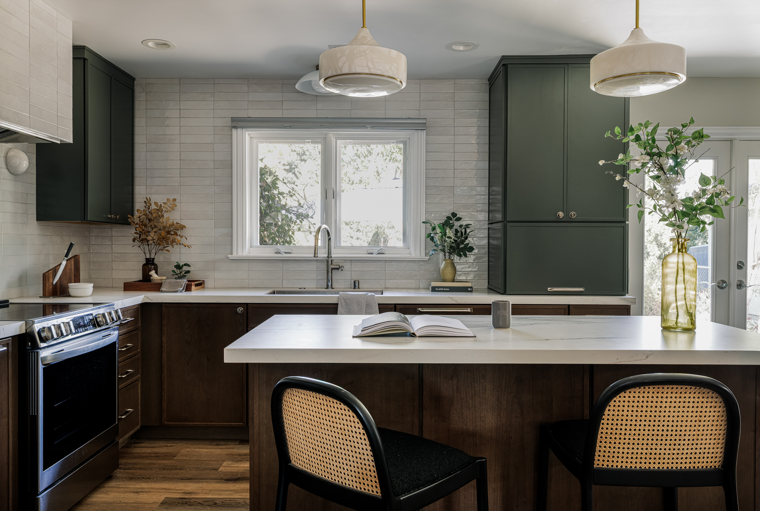 The finished kitchen with a marble-topped island and two cane stools, dark wood lower cabinets, a deep green pantry, a window above the sink and French doors bringing light in at right.