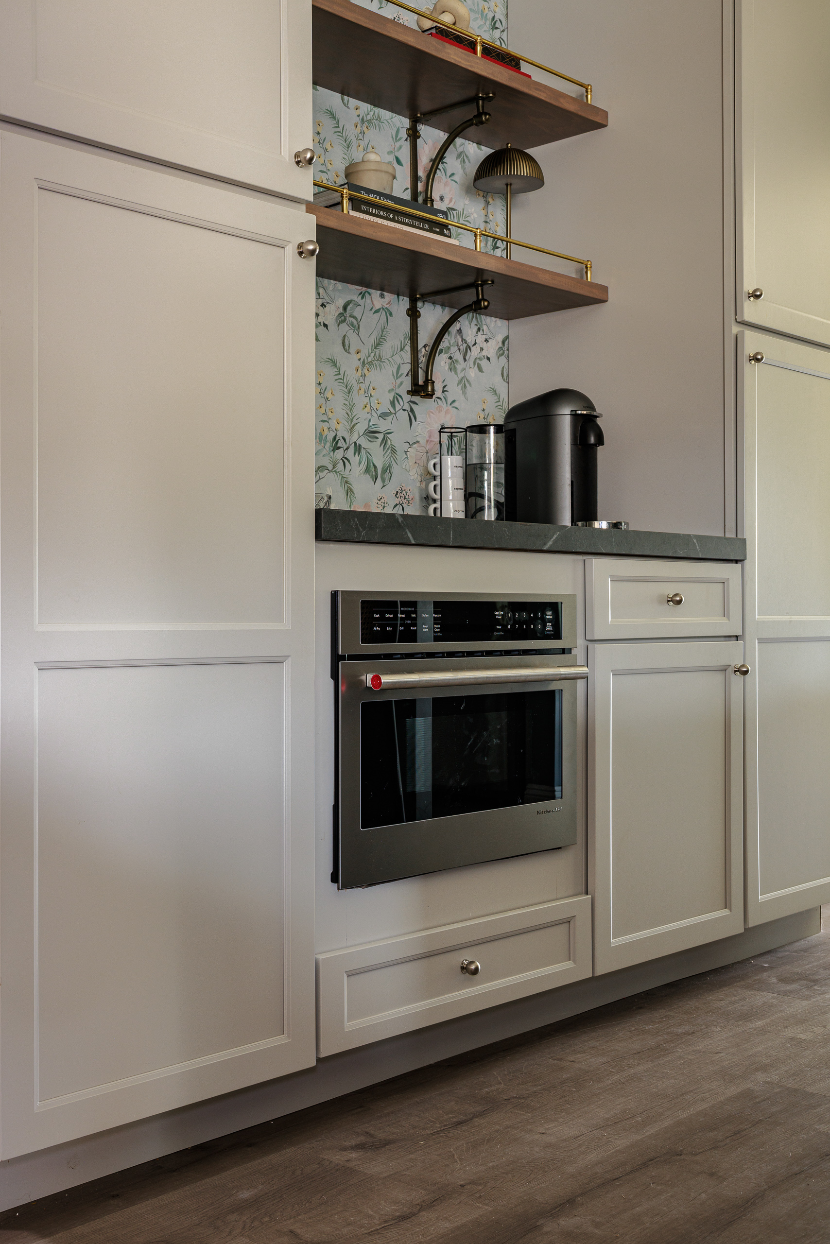 Built in wall microwave set into cream cabinetry below the coffee bar counter, with wallpaper and brass rail shelving above