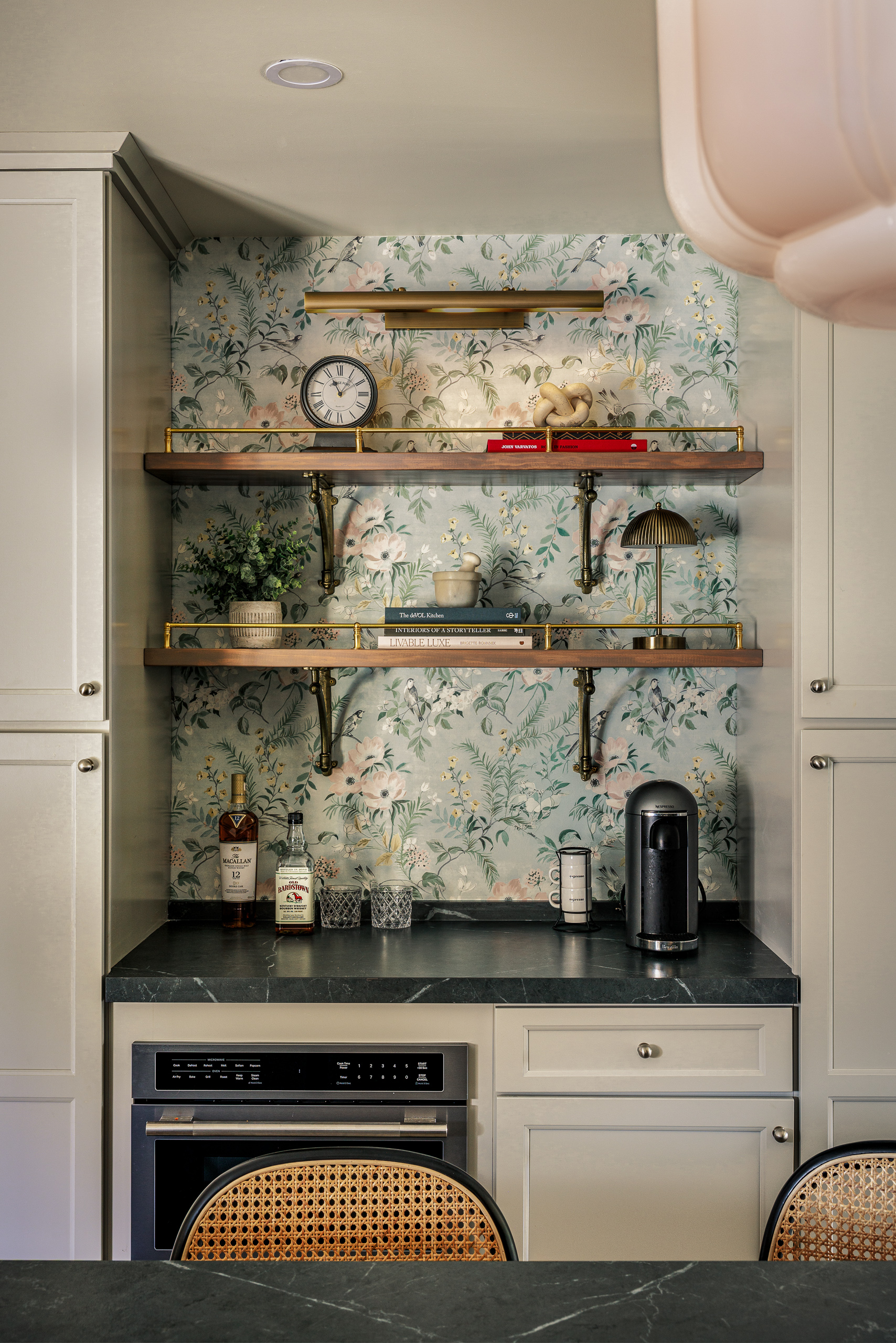 Floral bird wallpaper behind walnut shelves with brass gallery rails, a picture light above and glassware and books styled on the shelves