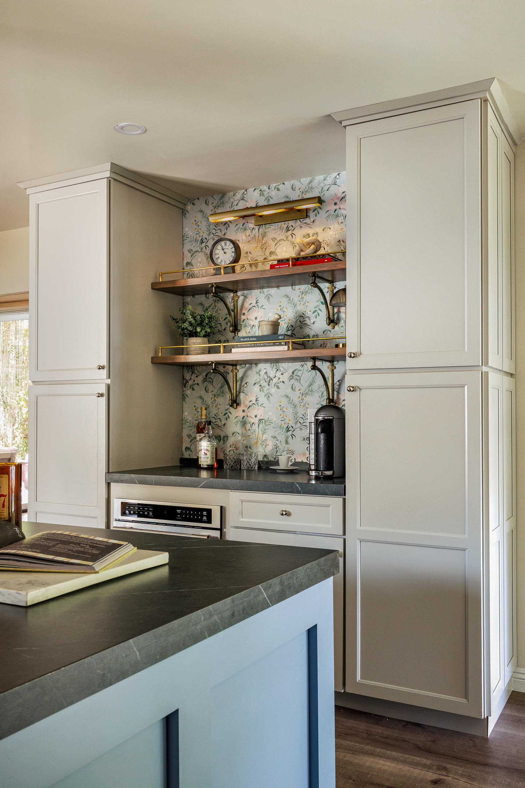 The coffee bar tucked between tall cabinets, with floral wallpaper, brass rail shelving and a soapstone counter beside the blue island