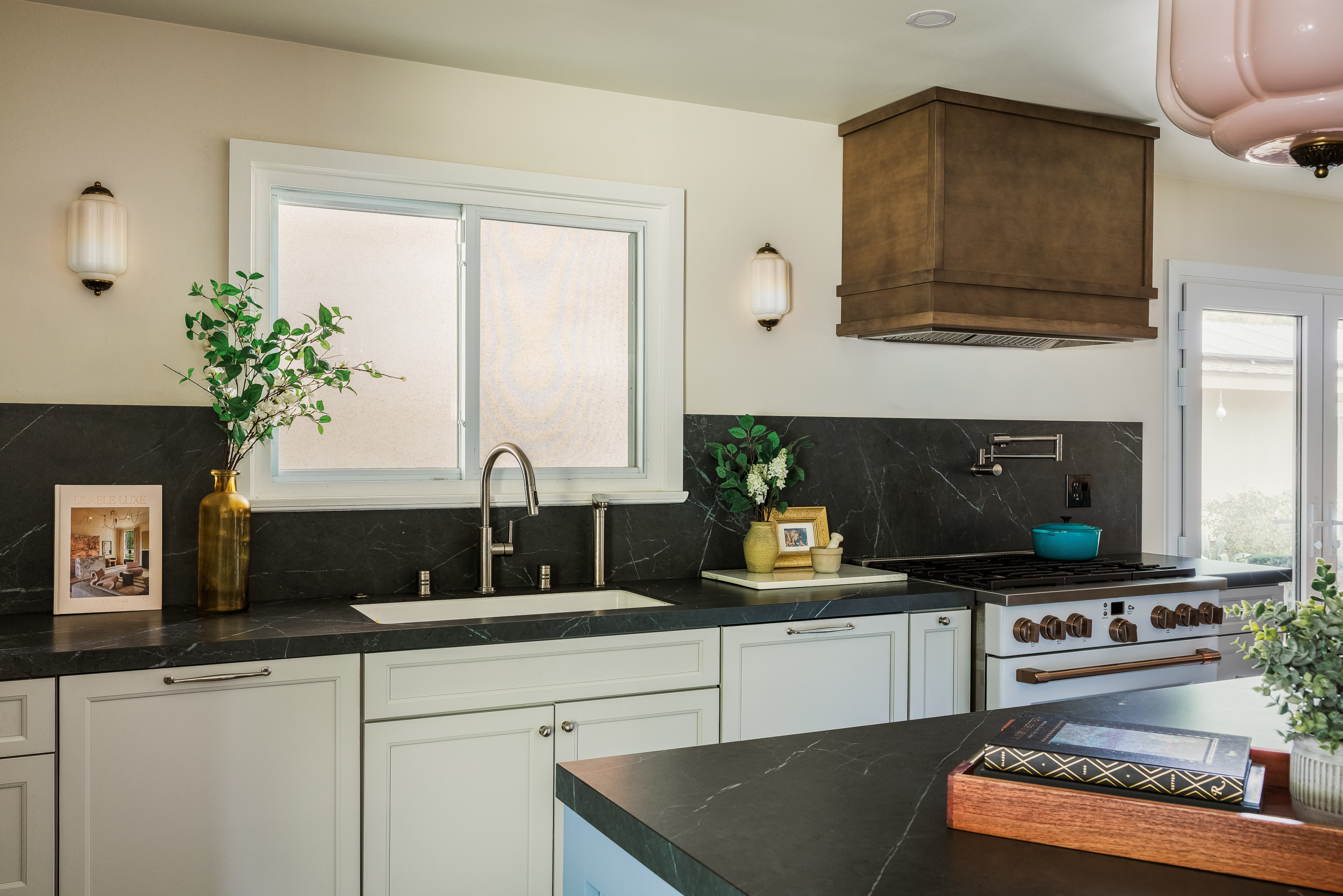 Undermount sink beneath a window with a full height soapstone backsplash, fluted glass sconces and a pot filler at the range