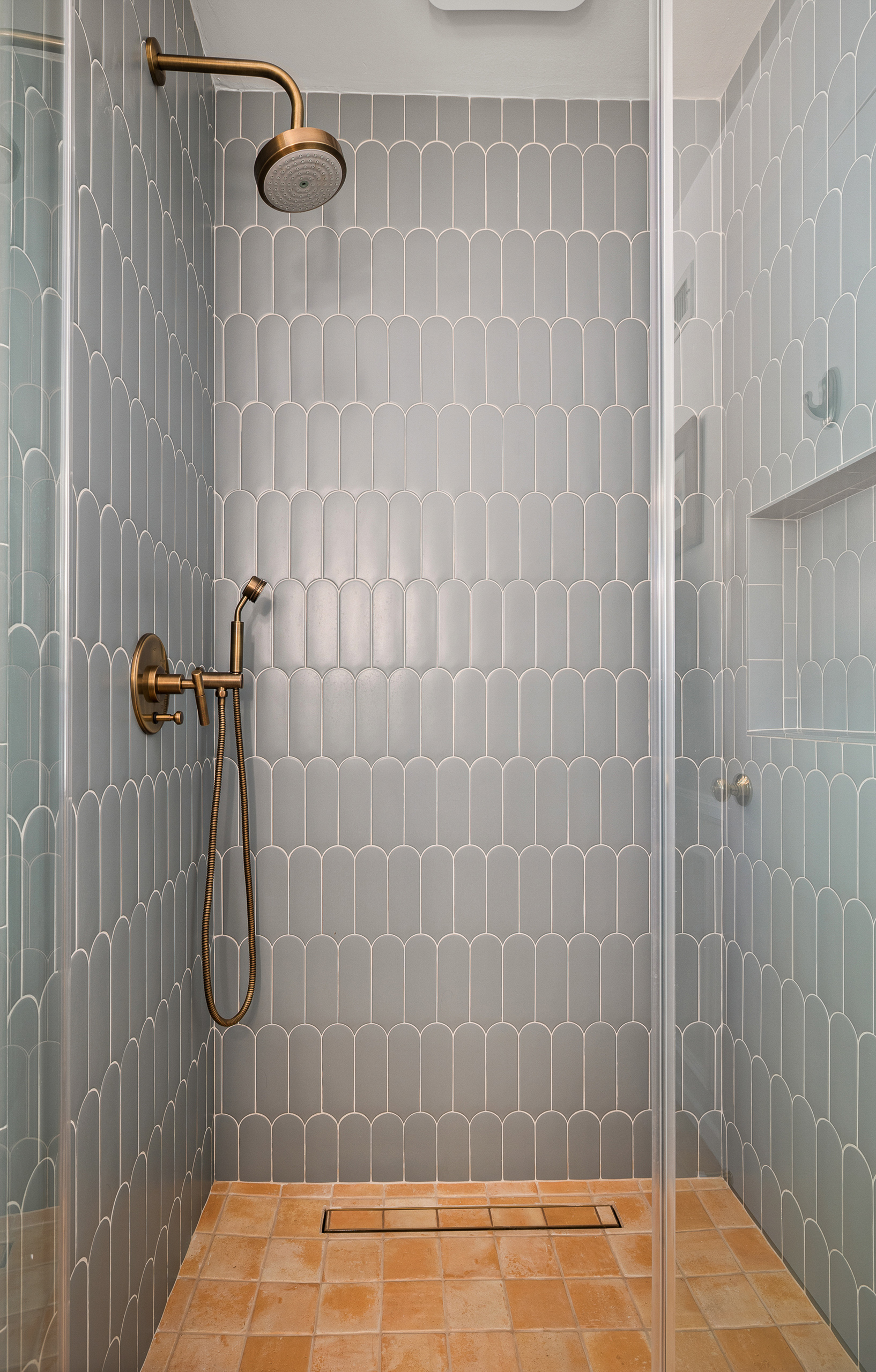 Walk-in shower lined in blue scalloped tile with an aged brass showerhead and handheld, over a terracotta tile floor