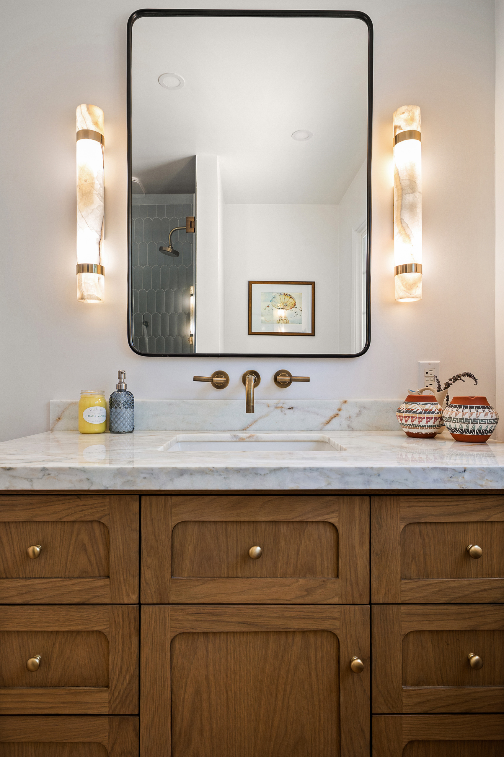 Wood vanity with a marble counter, wall mounted brass faucet, black framed mirror and a pair of alabaster sconces