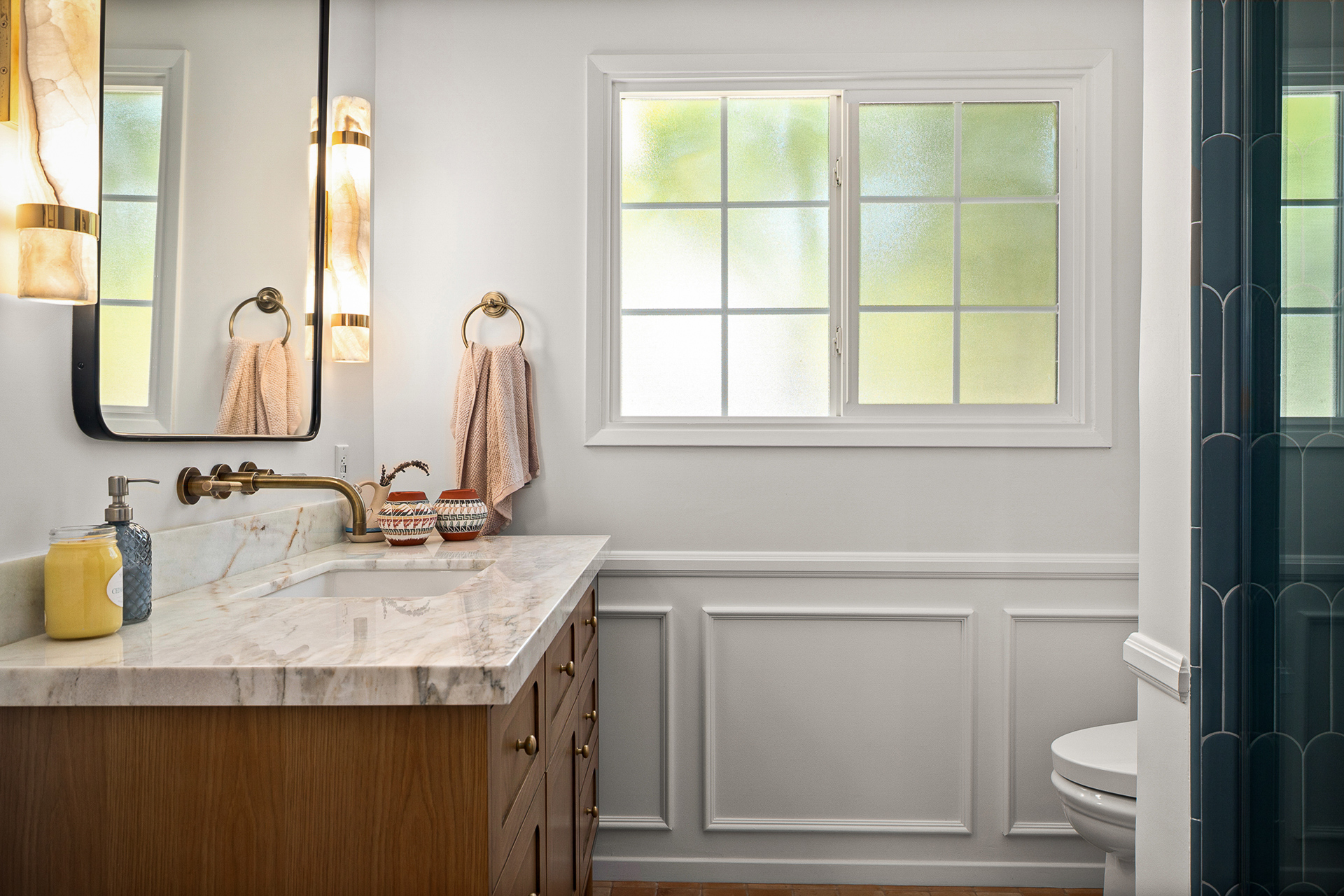 Bathroom with a wood vanity and marble counter, alabaster sconces, and a frosted window above white paneled wainscoting