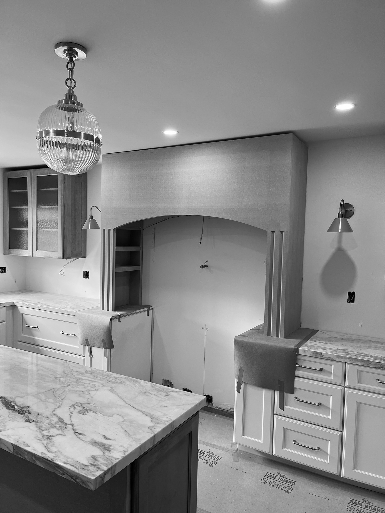The arched range hood surround framed and drywalled during construction, with marble counters set and a ribbed glass pendant already hung above the island