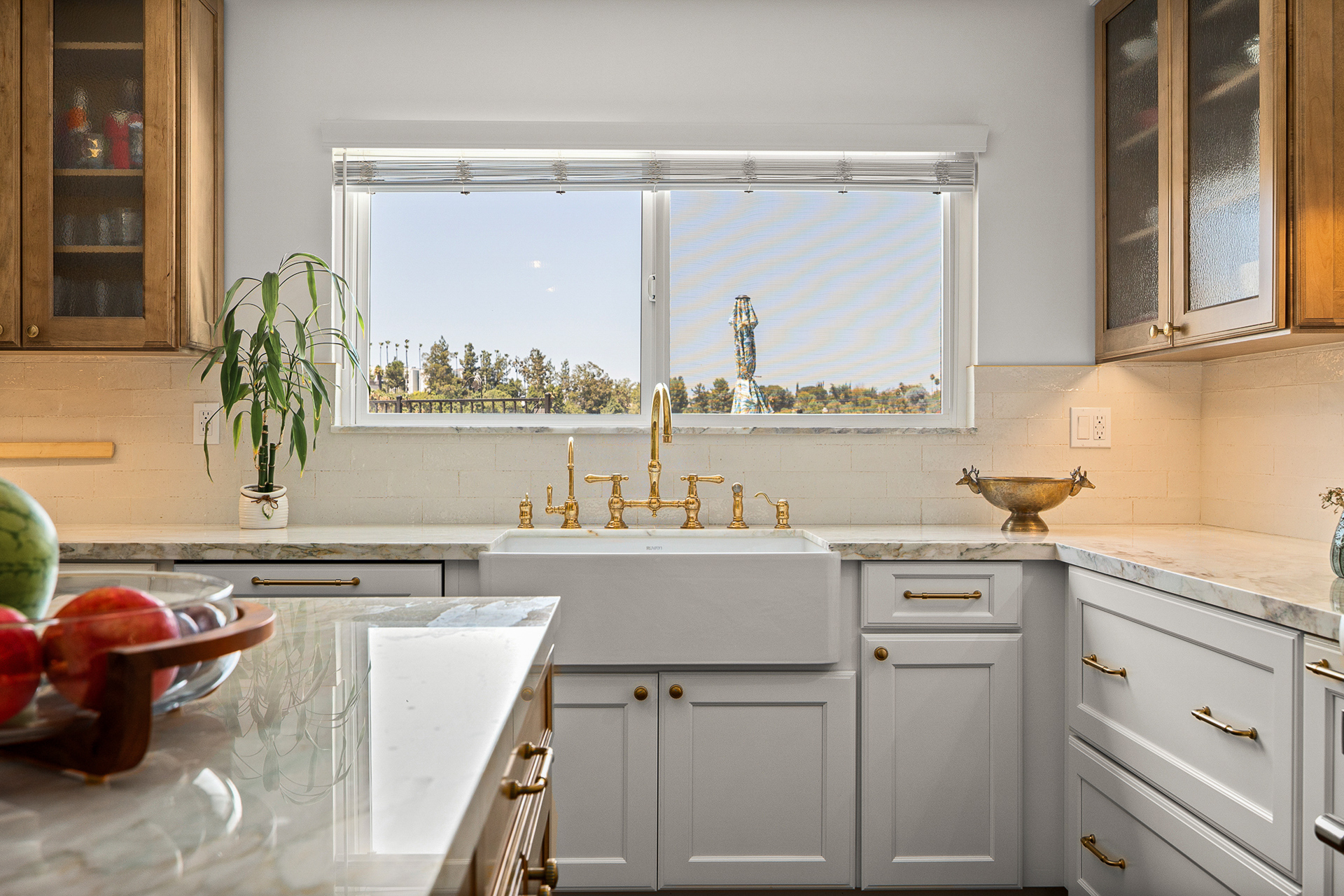 Farmhouse sink set beneath a window with an unlacquered brass bridge faucet, marble counters and a pale limestone backsplash